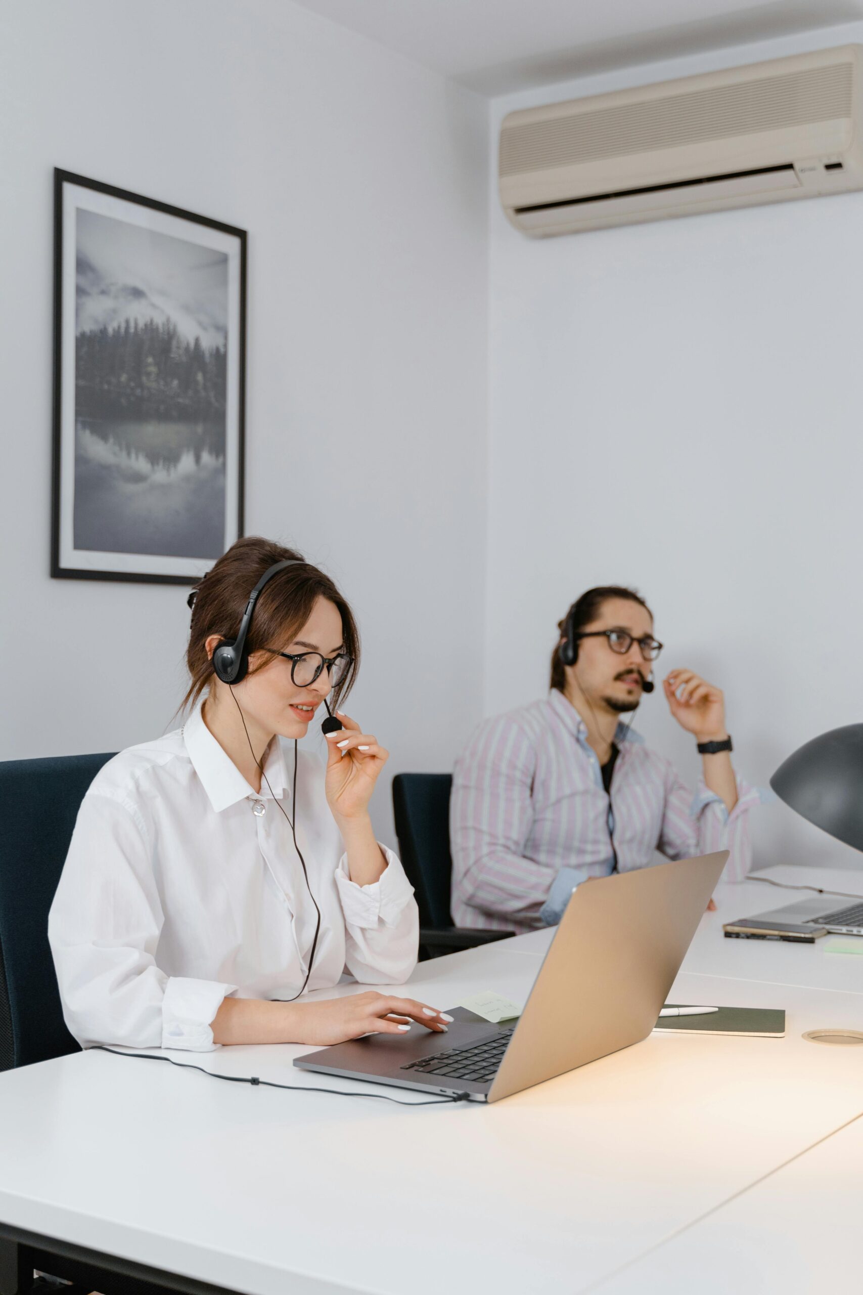 Two professionals working remotely in a modern office space, wearing headsets and using laptops.